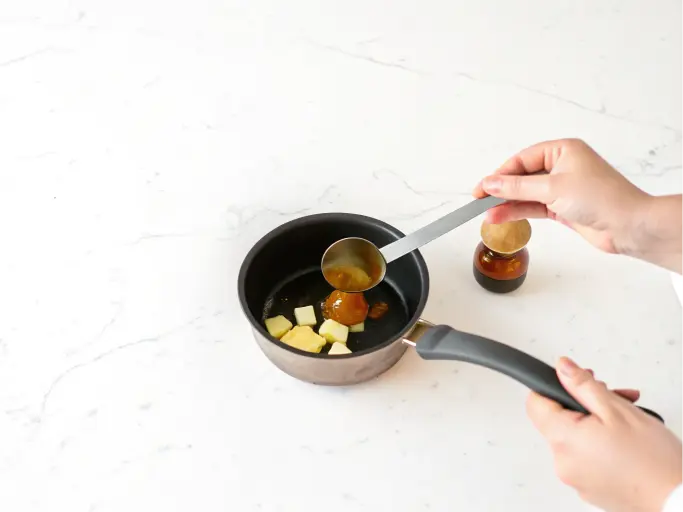 A hand is pouring honey into a saucepan containing other ingredients.